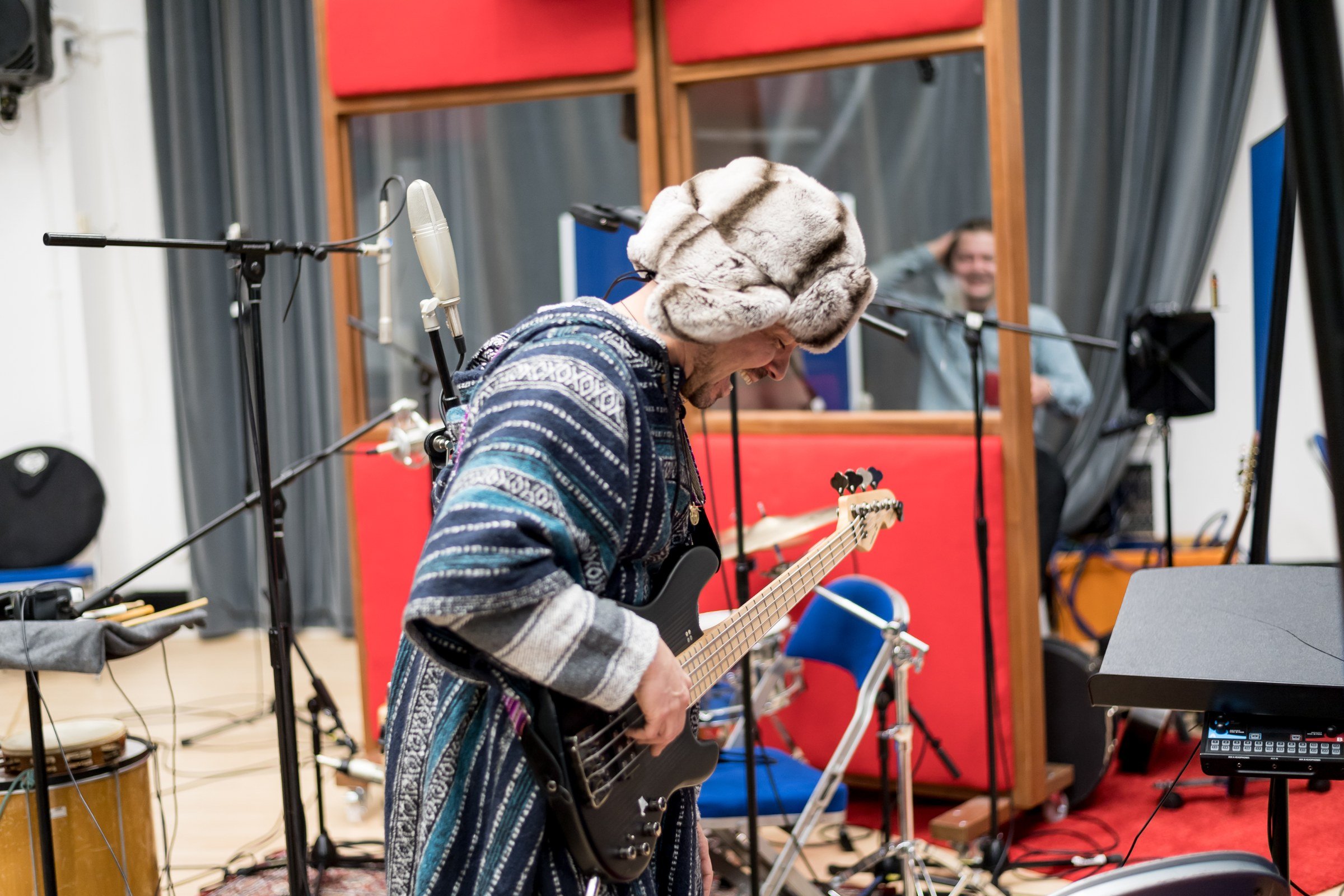 Stuart Zender playing bass in the live room, in a fur hat, screens and microphone stands around him