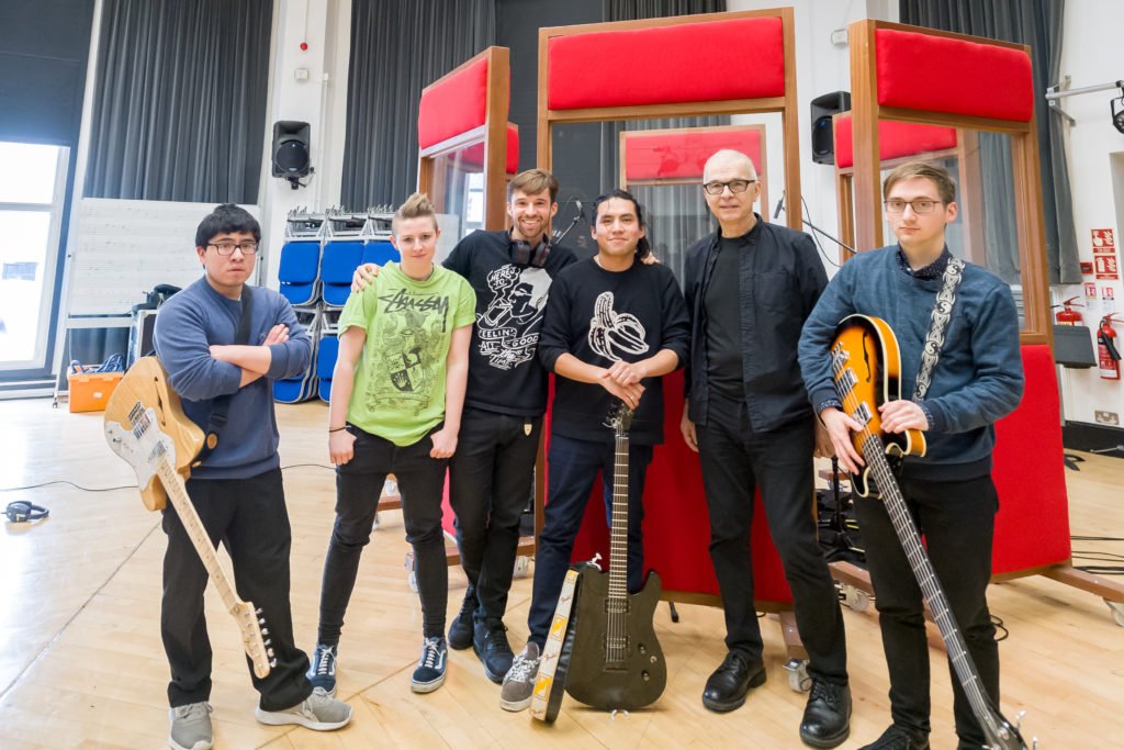 Tony Visconti pictured with Artbreak, who are holding their instruments, stood in front of red gobos in the studio live room