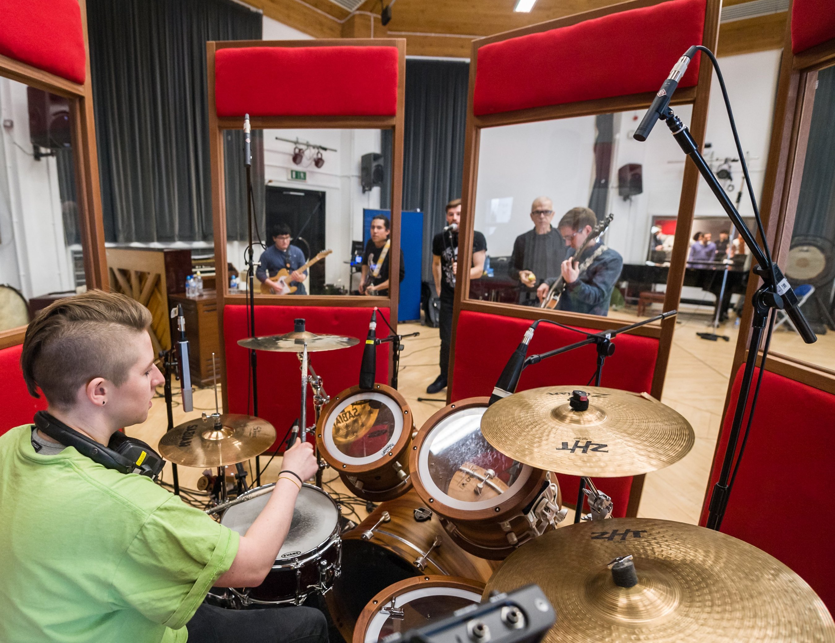 A drummer recording in the live room, close-miked behind red screens, with guitarists and a bassist playing beyond them