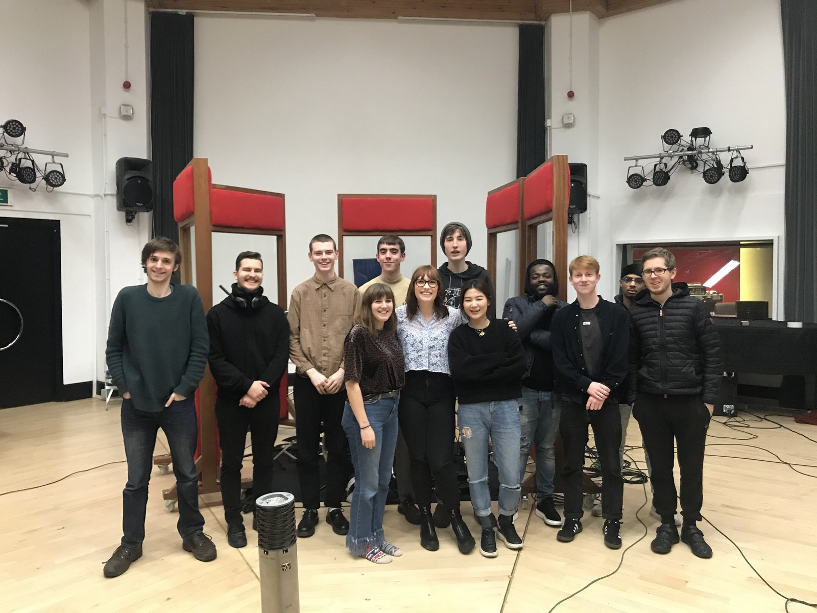 Thirteen music students standing together in the live room, the red booths and octagonal ceiling behind them