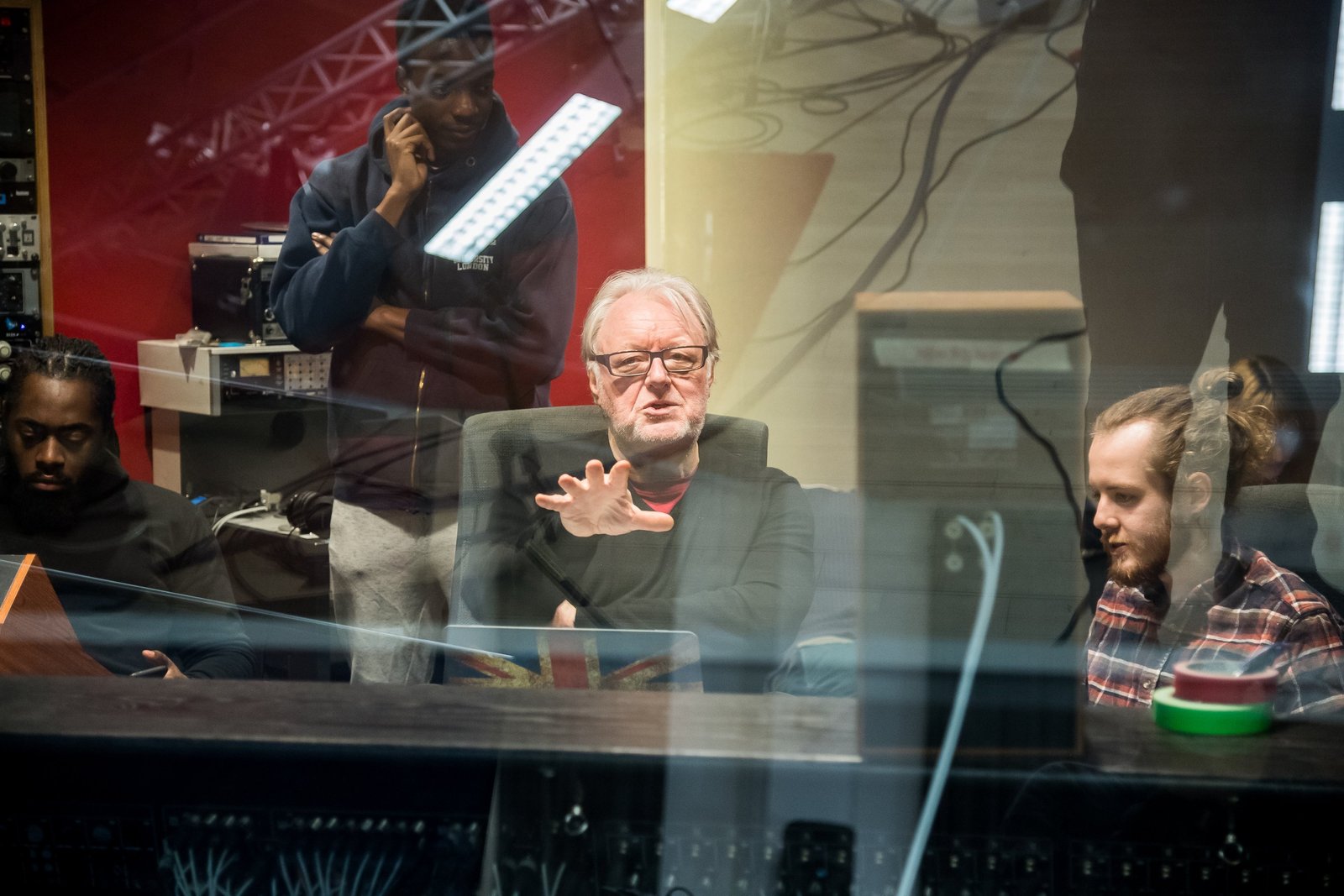 Chris Kimsey explaining a decision at the console, with three students gathered around him, seen through the control room glass